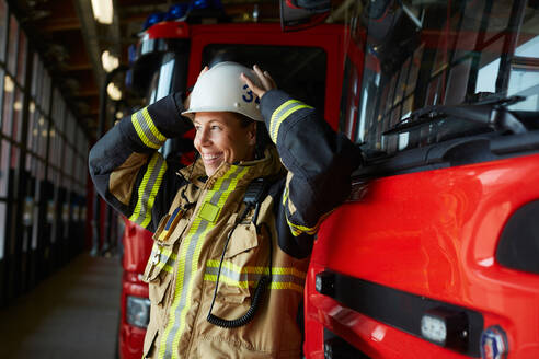 Lächelnder weiblicher Feuerwehrmann mit Helm, der neben einem Feuerwehrauto auf der Feuerwache steht - MASF14211