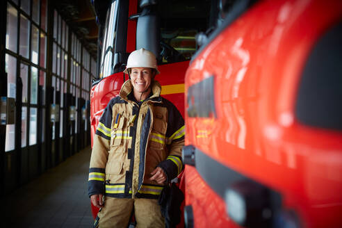 Porträt einer lächelnden weiblichen Feuerwehrfrau, die neben einem Feuerwehrauto auf der Feuerwache steht - MASF14213