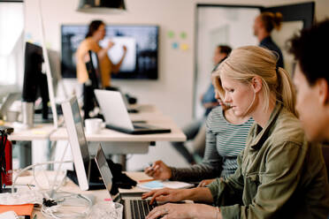Weibliche Führungskraft tippt auf einem Laptop, während sie neben Kollegen in einem kreativen Büro sitzt - MASF14236