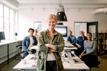 Happy businesswoman standing with arms crossed at desk with colleagues in background - MASF14251