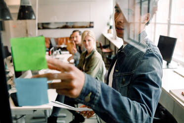 Businessman showing adhesive note to creative team seen from glass at workplace - MASF14264