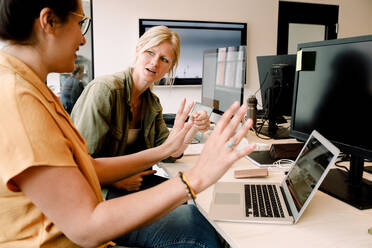 Female business professionals discussing over laptop while sitting at desk in office - MASF14283