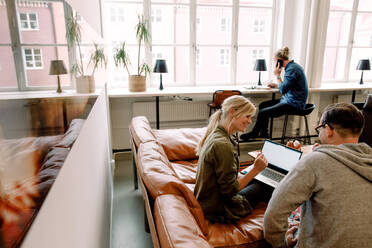 Coworkers discussing over laptop on sofa while businessman working in background at workplace - MASF14289