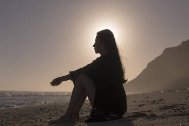 Seitenansicht der nachdenklichen Frau sitzt am Strand gegen den Himmel bei Sonnenuntergang - CAVF68356