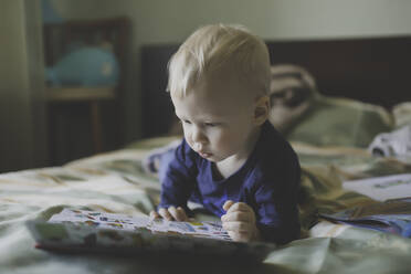 Junge schaut sich ein Buch an, während er auf dem Bett im Schlafzimmer liegt - CAVF68403