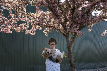 Porträt eines Jungen, der einen Hund trägt, während er an einem Baum vor Wellblech steht - CAVF68414