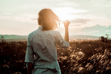 Rückansicht einer Frau, die ihr Haar hält, während sie auf einem Feld gegen den Himmel bei Sonnenuntergang steht - CAVF68423