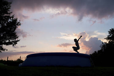 Unbekümmerter Junge mit erhobenen Armen, der in der Abenddämmerung gegen den bewölkten Himmel in ein Schwimmbecken springt - CAVF68442