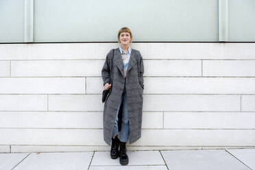 Portrait of strawberry blonde young woman wearing grey coat leaning against wall - FLLF00334