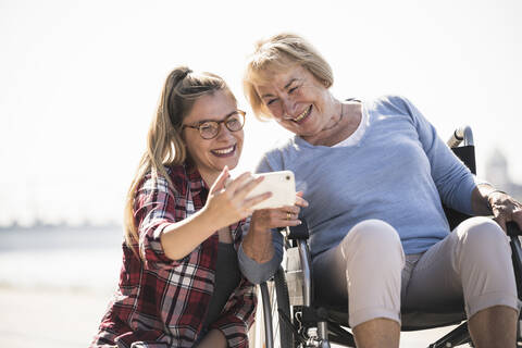 Junge Frau mit ihrer lächelnden Großmutter, die im Rollstuhl sitzt und ein Selfie macht, lizenzfreies Stockfoto