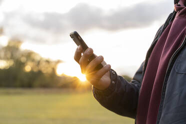 Mann steht in ländlicher Landschaft bei Sonnenuntergang und hält ein Smartphone - AFVF04162