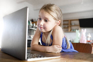 Porträt eines kleinen Mädchens, das sich zu Hause an den Küchentisch lehnt und auf den Laptop schaut - KMKF01133