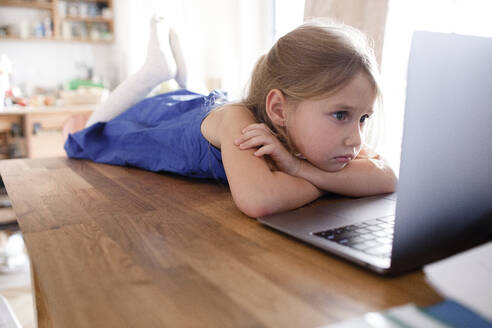 Portrait of sad little girl lying on kitchen table at home looking at laptop - KMKF01134