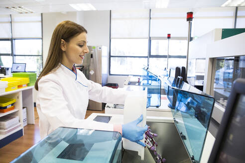 Side view of young woman in lab coat pouring vials of blood samples into refrigerator while working in research lab - ABZF02805