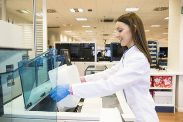 Side view of young woman in lab coat pouring vials of blood samples into refrigerator while working in research lab - ABZF02807