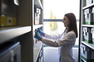 Side view of woman in white robe putting box with samples on shelf in lab storage - ABZF02809
