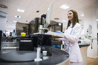 Young woman in white apparel using sample analyzer while working in research lab - ABZF02813