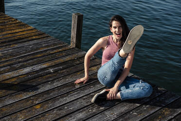 Portrait of screaming young woman sitting on jetty kicking in the air, Lake Starnberg, Germany - WFF00154