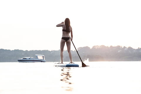 Rückenansicht einer jungen Frau, die bei Sonnenuntergang Stand-Up-Paddle-Boarding betreibt, Starnberger See, Deutschland, lizenzfreies Stockfoto