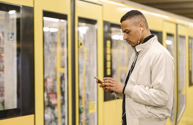 Mann steht auf dem Bahnsteig einer U-Bahn-Station und benutzt Kopfhörer und Mobiltelefon, Berlin, Deutschland - AHSF01088