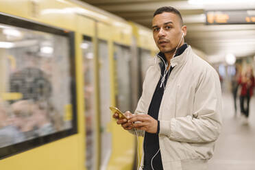 Porträt eines Mannes auf dem Bahnsteig einer U-Bahn-Station, der Kopfhörer und ein Mobiltelefon benutzt, Berlin, Deutschland - AHSF01089