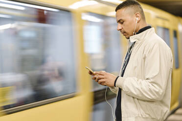 Mann steht auf dem Bahnsteig einer U-Bahn-Station und benutzt Kopfhörer und Mobiltelefon, Berlin, Deutschland - AHSF01090