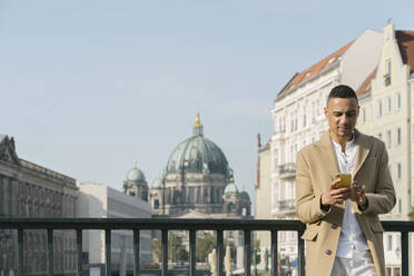 Porträt eines Geschäftsmannes, der auf einer Brücke in der Stadt steht und auf sein Handy schaut, Berlin, Deutschland - AHSF01095