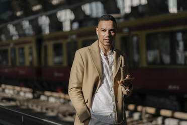 Porträt eines Mannes, der am Bahnhof steht und ein Smartphone und Kopfhörer benutzt, Berlin, Deutschland - AHSF01100