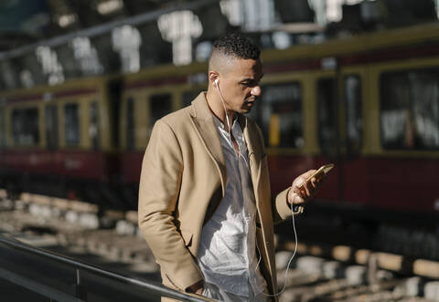 Mann steht am Bahnhof und benutzt Smartphone und Kopfhörer, Berlin, Deutschland, lizenzfreies Stockfoto