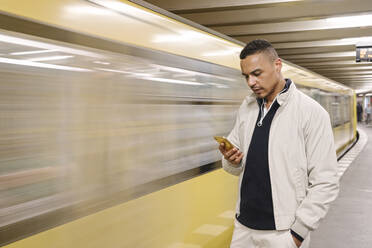 Porträt eines Mannes, der vor einem fahrenden U-Bahn-Zug steht und auf sein Handy schaut, Berlin, Deutschland - AHSF01125