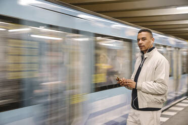 Portrait of man with cell phone and earphones standing in front of driving underground train, Berlin, Germany - AHSF01126