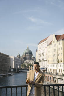 Portrait of businessman standing on a bridge looking at smartphone, Berlin, Germany - AHSF01128