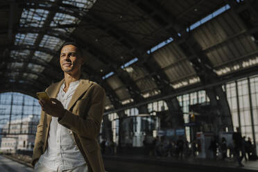 Portrait of man standing at train station with smartphone looking at distance, Berlin, Germany - AHSF01136
