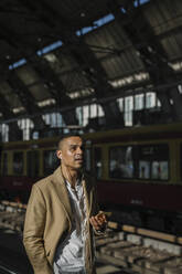 Porträt eines Geschäftsmannes, der am Bahnhof steht und ein Smartphone und Kopfhörer benutzt, Berlin, Deutschland - AHSF01137