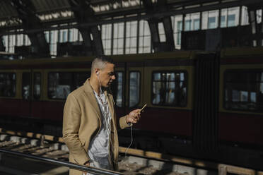 Mann steht am Bahnhof und benutzt Smartphone und Kopfhörer, Berlin, Deutschland - AHSF01138