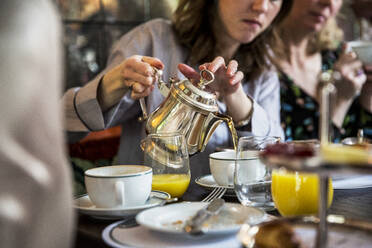 Close up of woman sitting at a table, pouring tea from silver tea pot. - MINF12687