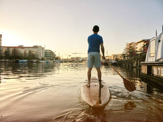 Mann steht auf einem Paddelbrett auf einem Fluss in der Morgendämmerung, Aufnahme von hinten - MINF12792