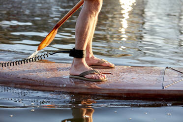 Nahaufnahme der Beine eines Mannes, der auf einem Paddelbrett auf einem Fluss steht - MINF12798