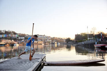 Mann mit Paddel, der sich die Beine am Ufer des Flusses ausstreckt, bevor er sein Paddelbrett benutzt - MINF12803