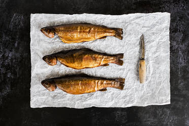 High angle close up of three freshly smoked whole trout and a knife on a white paper. - MINF12867