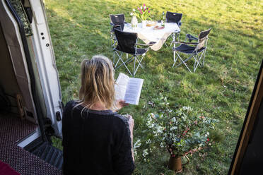 Hochformatige Rückansicht einer Frau, die in der Tür eines auf einer Wiese geparkten Wohnmobils sitzt und ein Buch liest. - MINF12923
