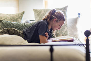 13 year old girl lying in her bed reading by window light - MINF12959
