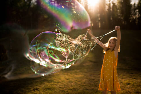 9 year old girl making giant bubble in summer light - CAVF68508