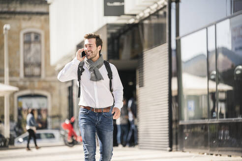 Lächelnder junger Mann mit Rucksack beim Telefonieren in der Stadt - UUF19575