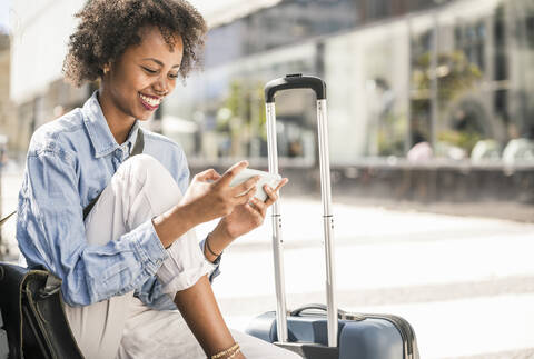Happy young woman sitting on a bench with suitcase using cell phone stock photo