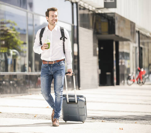 Lächelnder junger Mann mit Rollkoffer in der Stadt unterwegs, lizenzfreies Stockfoto