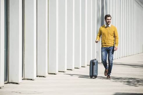 Young man with rolling suitcase and earphones in the city on the go - UUF19613