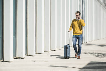 Smiling young man with rolling suitcase and earphones in the city on the go - UUF19614