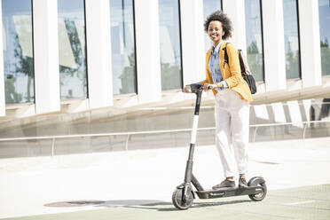 Happy young woman with earphones riding e-scooter in the city - UUF19619