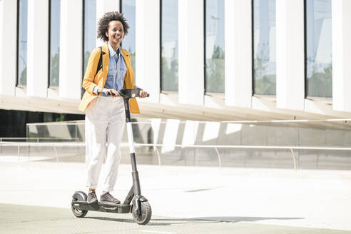 Happy young woman with earphones riding e-scooter in the city - UUF19620